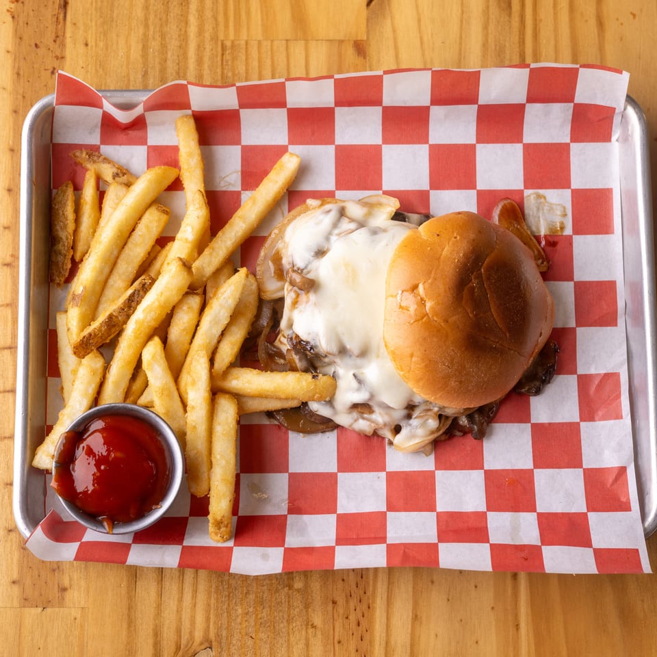 Cheeseburger with melted cheese, caramelized onions, and mushrooms, served with fries and ketchup on a checkered tray.