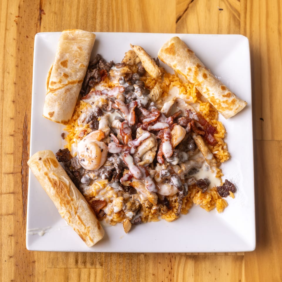 Plate of rice, beef, chicken, shrimp, and bacon with creamy sauce, served with tortillas.