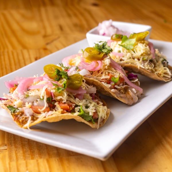 Three tacos topped with shredded cheese, pickled red onions, jalapeños, and cilantro on a white plate.