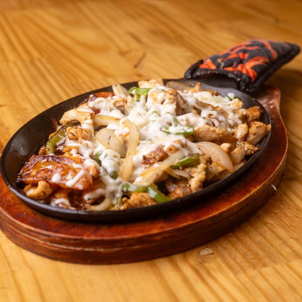 Chicken fajitas with melted white cheese, onions, and green peppers in a hot skillet on a wooden table.