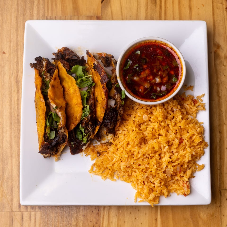 Three birria tacos with rice and a side of consommé on a white plate.