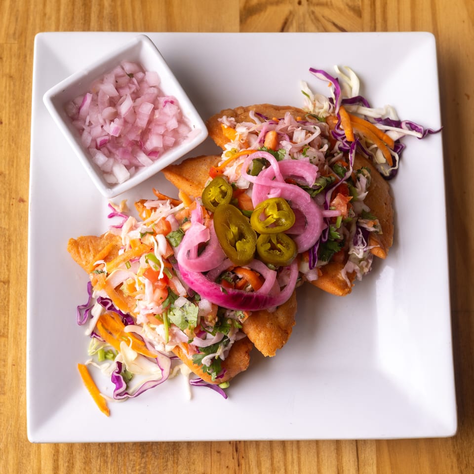 Golden fried tacos topped with colorful cabbage slaw, pickled onions, and jalapeños, with a side of diced red onions.