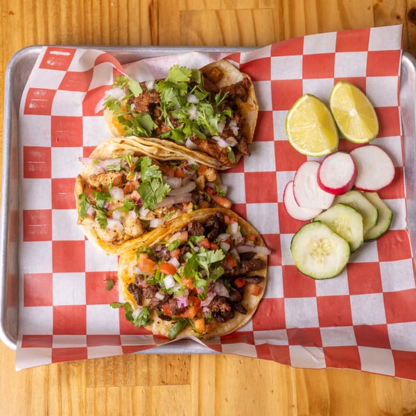 Three vibrant tacos with various toppings, lime, radishes, and cucumbers on a metal tray with checkered paper.