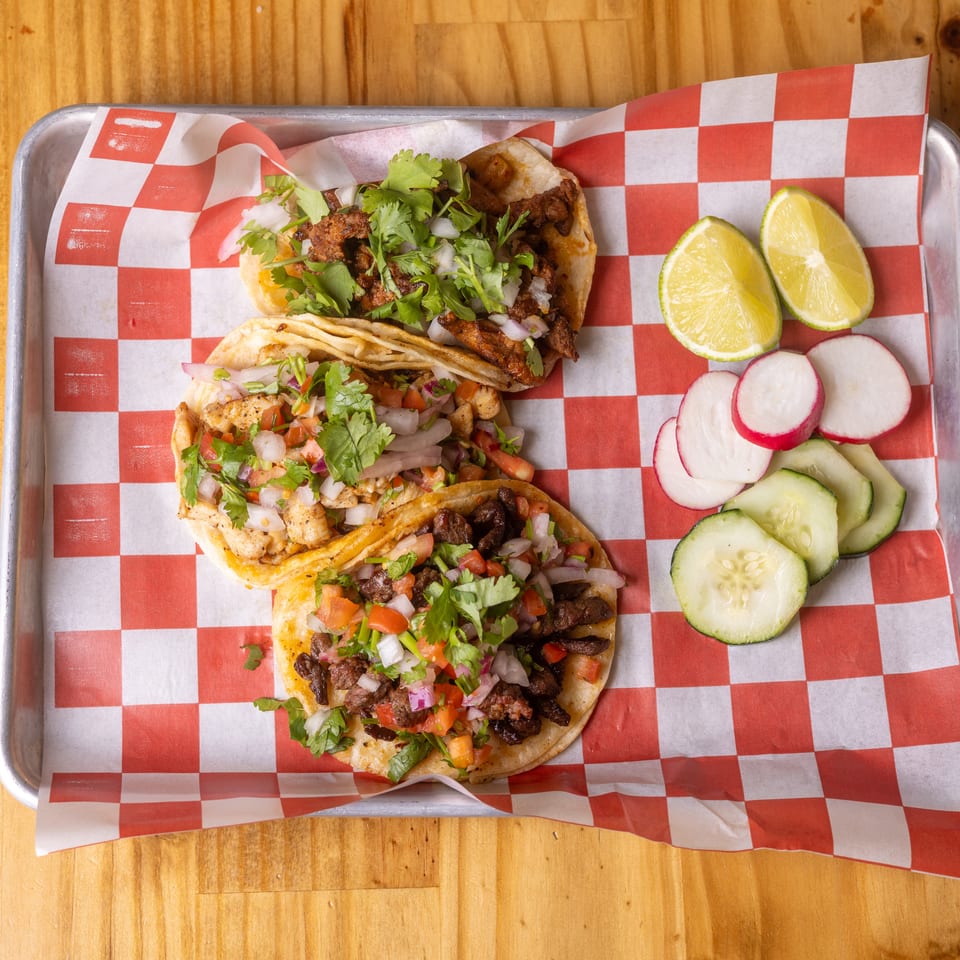Three vibrant tacos with various toppings, lime, radishes, and cucumbers on a metal tray with checkered paper.