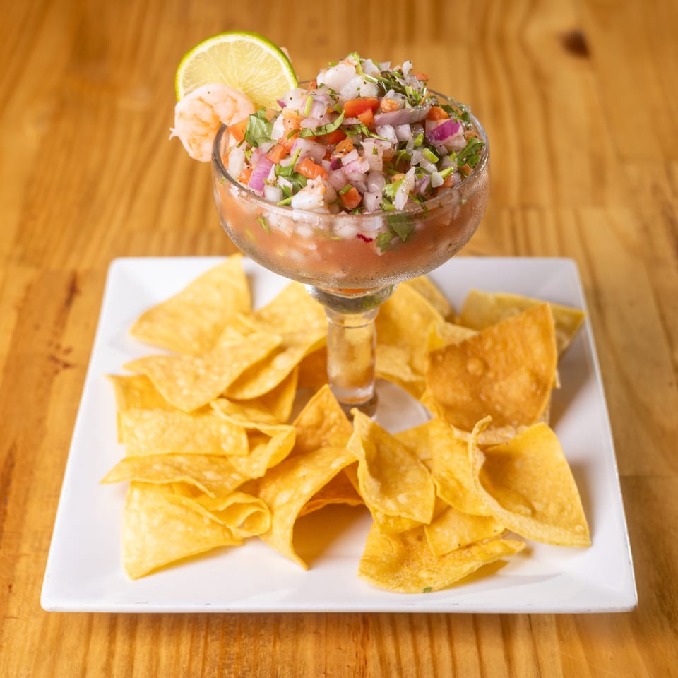 A glass of shrimp ceviche with diced onions, tomatoes, cilantro, lime, and tortilla chips.