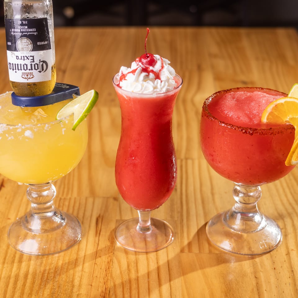 Three colorful drinks: a yellow margarita with a Corona, a strawberry frozen drink, and a chili-rimmed red frozen drink.