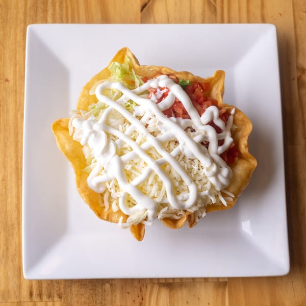 Taco salad in a fried tortilla bowl with lettuce, tomatoes, cheese, and sour cream on a white plate.